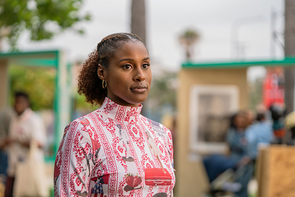 A woman with braided hair and gold hoop earrings wearing a red and white patterned high-neck top.