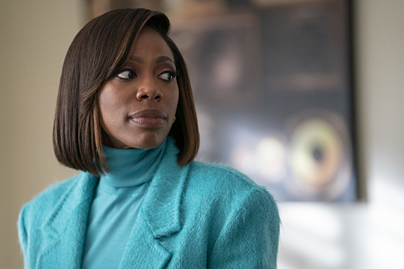 A woman with short dark hair wearing a turquoise turtleneck and textured coat looks off-camera.