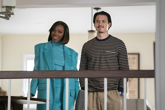 A woman in a teal blazer and turtleneck stands next to a man in a navy and gold striped long-sleeve shirt.