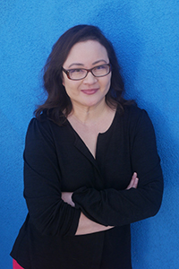 A woman with dark hair and glasses wearing a black shirt, standing with arms crossed against a blue wall.