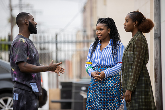 A man in a tie-dye shirt gestures while talking to two women standing on a sidewalk.