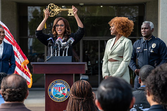 A woman in a black blazer speaks at a podium with microphones, flanked by others outdoors.