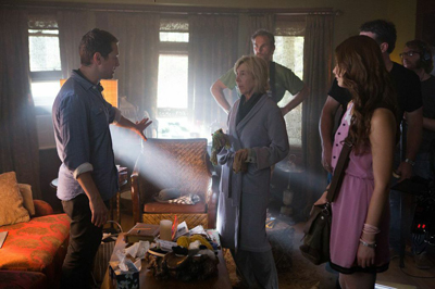 A man in a blue shirt gestures toward a woman in a grey robe while others stand nearby in a dimly lit room.