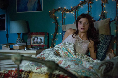 A young woman with long dark hair sits up in a bed with a surprised look, surrounded by fairy lights.