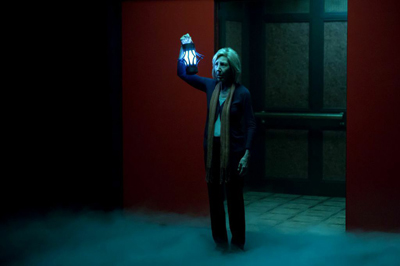 A woman in a dark blue sweater holds up a glowing blue lantern amidst low-lying fog.