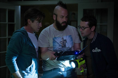 Three men gather closely in a dimly lit room to examine a small electronic device held by the man in the center.