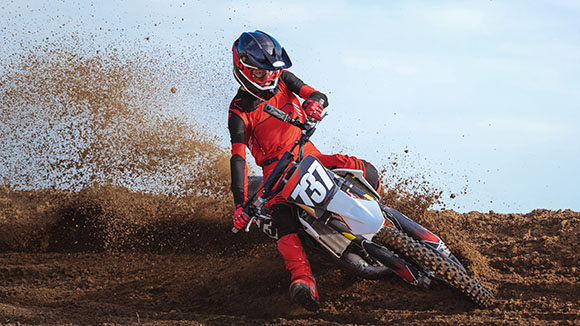A motocross rider in red and black gear leans into a turn on a dirt track, kicking up a large spray of soil.