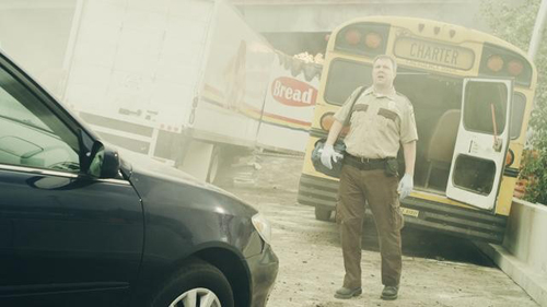 A man in a tan uniform stands between a black car and a yellow bus near a white bread truck.