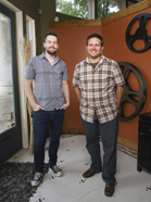 Two smiling men standing indoors next to a wall decorated with large film reels.