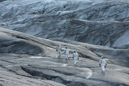 Four people in white protective suits walking across a vast, grey, textured glacier.