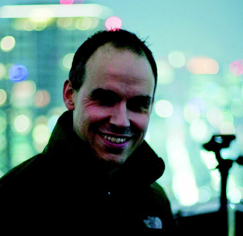 A man with short hair smiles toward the camera against a bokeh city background.