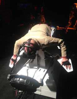 Person wearing a VR headset and gloves, suspended horizontally over a machine with wires.