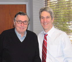 Two men, one in a black sweater and glasses, the other in a white shirt and red striped tie, smiling.