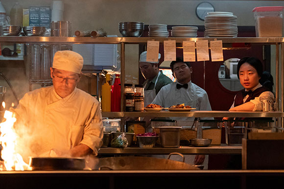 Chefs prepare food in a professional kitchen with flames rising from a pan in the foreground.