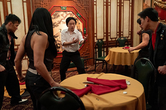 A man in a white shirt gestures toward a group of people around yellow-clothed tables in a decorated room.