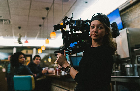 A woman in a black shirt holds a large professional video camera in a dimly lit cafe.