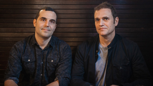 Two men sitting side by side in front of a dark wood-paneled wall.