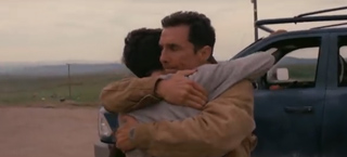 Two men hug tightly next to a blue car in a rural landscape.