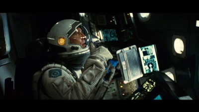 An astronaut in a white spacesuit and helmet sits in a dark spacecraft cockpit filled with glowing screens.
