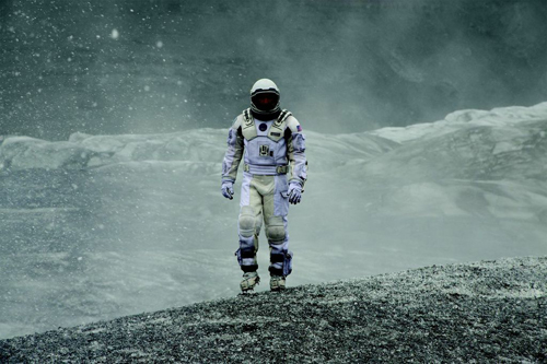 An astronaut in a white spacesuit stands on a rocky slope amidst falling snow and a misty gray landscape.