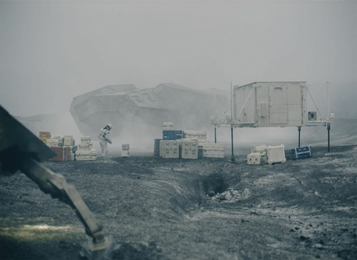 An astronaut stands near crates and a modular research station in a foggy, rocky landscape.
