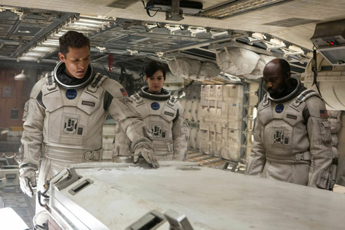 Three astronauts in white spacesuits stand around a console inside a spacecraft.
