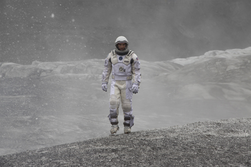 An astronaut in a white spacesuit stands amidst swirling dust on a rocky, grey planetary surface.