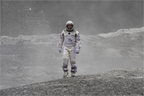 An astronaut in a white spacesuit stands on a rocky, dusty terrain under a hazy sky.