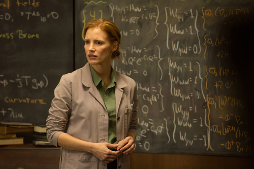 A woman with red hair wearing a light grey lab coat stands before a chalkboard filled with complex equations.