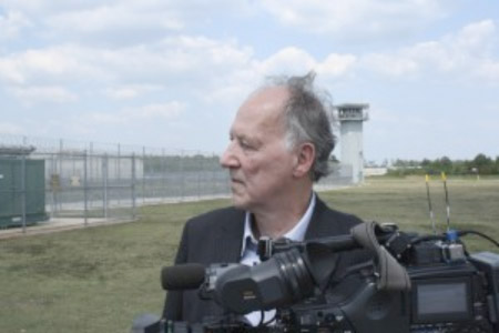 A man in a dark suit looks off-camera behind a professional video camera in an outdoor field.
