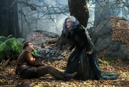 A man sits on a leaf-covered forest floor looking up at a woman in a dark, textured dress.