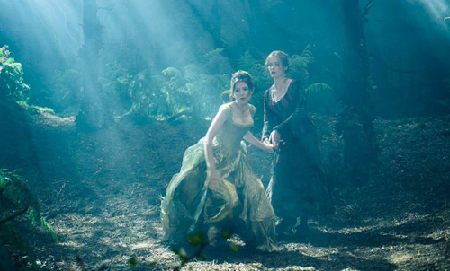 Two women in long dresses walk through a sun-dappled, misty forest.