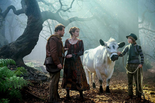 A man and woman in period clothing talk to a child holding a rope attached to a white cow in a misty forest.