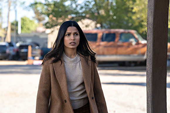 A woman with long dark hair wearing a tan sweater and brown coat looks concerned outdoors.
