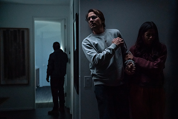 A man in a grey sweatshirt and a woman in red stand in a dark hallway near a silhouette in a doorway.