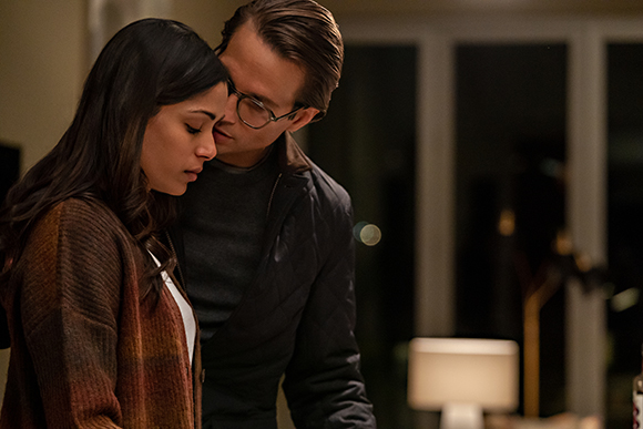 A man with glasses leans toward a woman with long dark hair in a low-light setting.