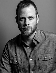 Black and white studio portrait of a man with short hair and a beard wearing a button-down shirt.