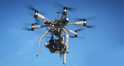 A large grey hexacopter drone with a professional camera gimbal flying against a clear blue sky.