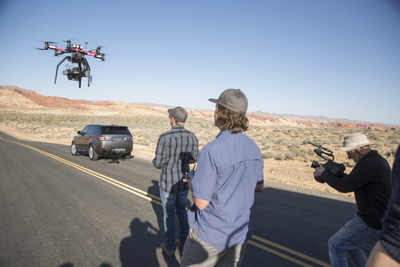 A drone with a camera flies over a road near a car while three people film in a desert landscape.