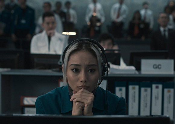 A woman with blonde hair and a headset stares intensely at a screen in a dark office.