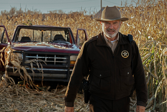 A smiling older man in a brown sheriff's uniform and hat stands in a cornfield near a damaged red truck.