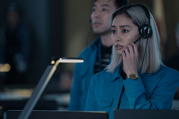A woman with blonde hair and a headset looks forward while wearing a blue jacket in a dark office setting.