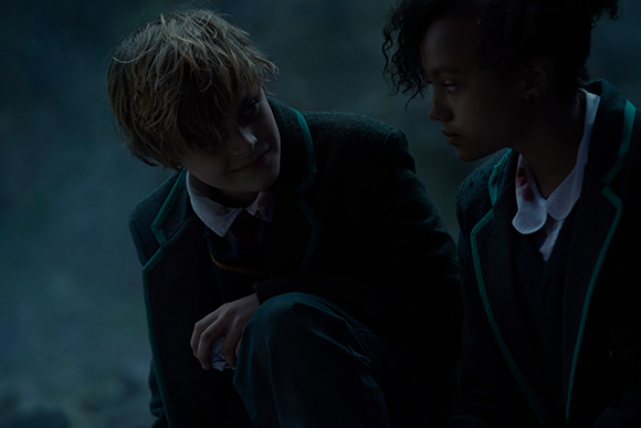 Two youths in dark school uniforms with teal trim sit close together in dim lighting.