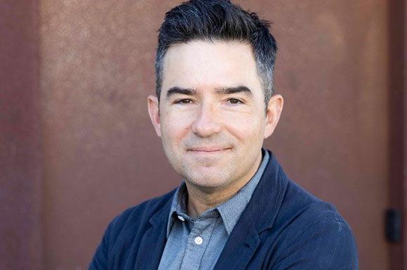 A man with short dark hair and a slight smile wearing a grey shirt and dark blazer.