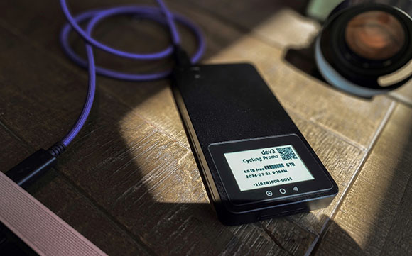 A black power bank with a purple cable and a white label sits on a wooden table next to a camera lens.