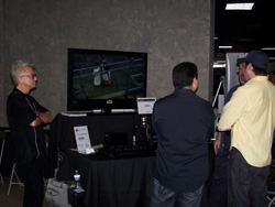 Several people stand around a display table with a television screen showing a video. One person in black stands with arms cr