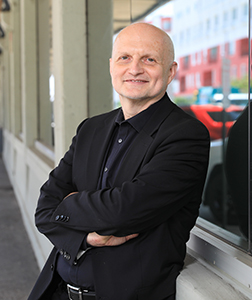 A bald man wearing a black blazer and button-down shirt smiles while leaning against a window ledge.