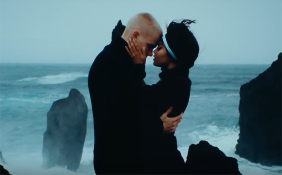 A man and woman in dark clothing embrace closely against a backdrop of a stormy ocean and dark rocks.