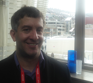 A man with short, dark hair and a light complexion smiles. He wears a purple shirt and a dark jacket. A red lanyard with 'UTA