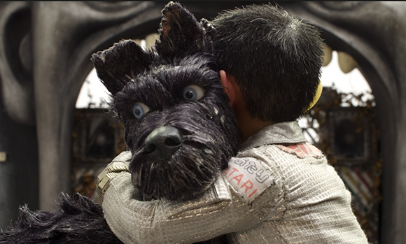 A person in a silver jacket hugs a large, black, textured puppet dog with blue eyes.